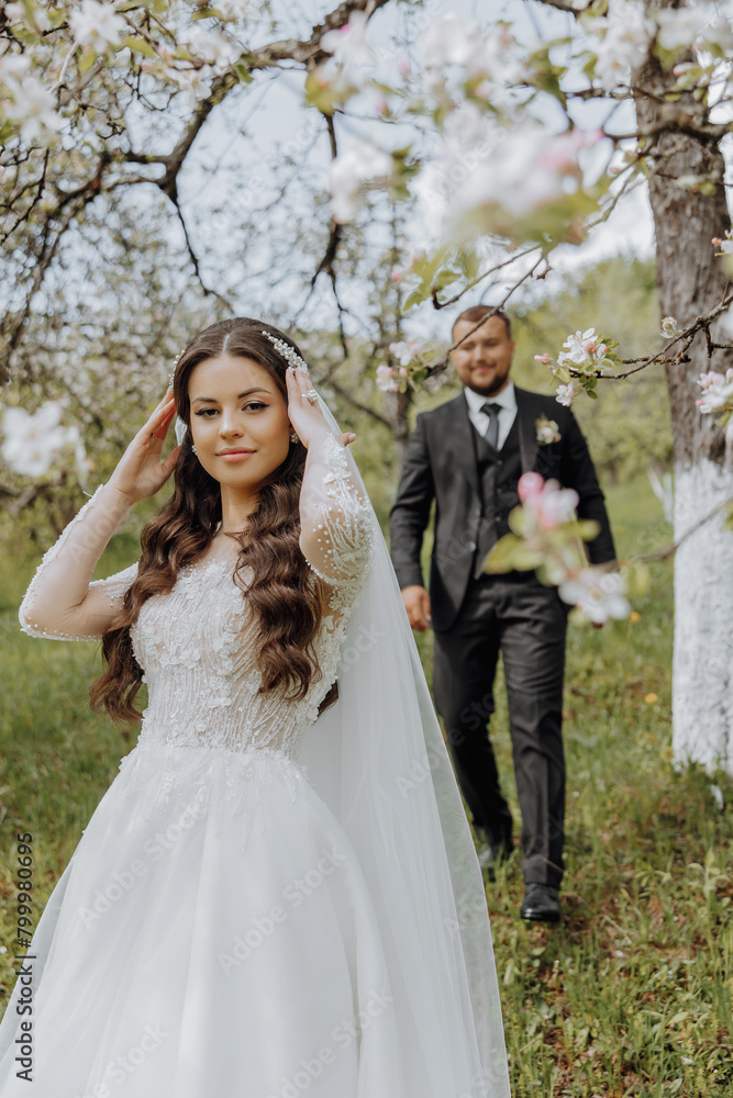 the first meeting of the bride and groom in a blooming garden. The bride in an incredible white dress with expensive hair and makeup. The groom in a black suit goes to the bride the first meeting of the bride and groom in a blooming garden. The bride in an incredible white dress with expensive hair and makeup. The groom in a black suit goes to the bride