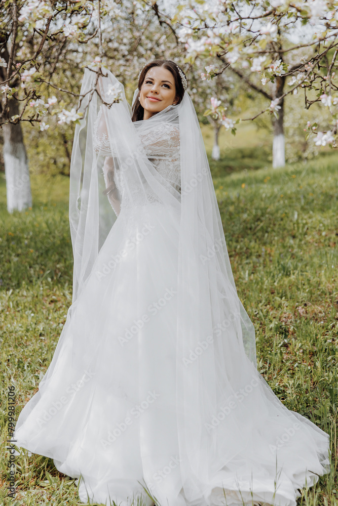 portrait of a very beautiful bride with long curly hair and wearing a white dress and long veil in a blooming spring garden. Naarechena holds her veil with her hands on a green background of grass