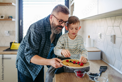 Boy filling pancake with fruit, sweets. Father spending time with son at home, making snack together, cooking. Fathers day concept. photo