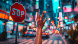 A male hand is seen reaching for a red stop sign with blurred city lights and urban architecture