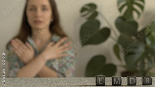 Letters EMDR written on black stone cubes. A female is touching and tapping her shoulders in a blurred background. Eye Movement Desensitization and Reprocessing Psychotherapy Treatment concept.