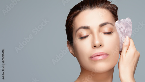 Woman practicing facial massage with gua sha stone, depicting beauty and skin care for anti-aging and spa treatment, isolated on gray background