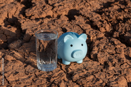 Heat-cracked clay in the desert, a glass of drinking water and a piggy bank