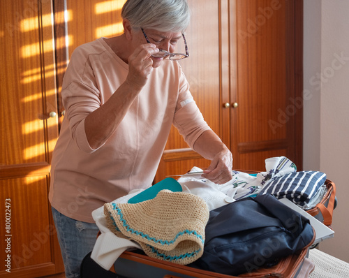 A smiling mature lady packs her suitcase for the trip to Paris to see the Olympics. Happy old woman in bedroom excited for the trip organizes her luggage essential for stay in hotel © luciano