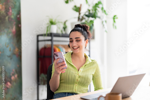 A jovial young woman with looks at her smartphone, laughing in a bright office space with stylish decor. photo