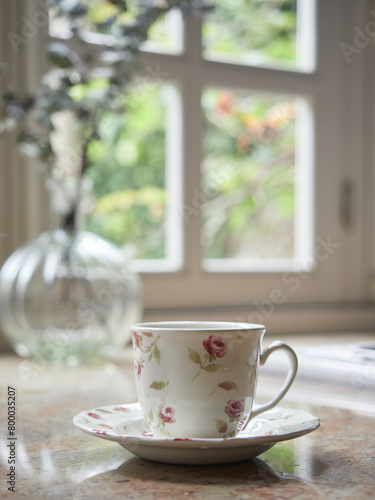 close-up view porcelain tea cup and saucer on kitchen countertop  background out of focus window and glass vase. Cozy cottage indoors.