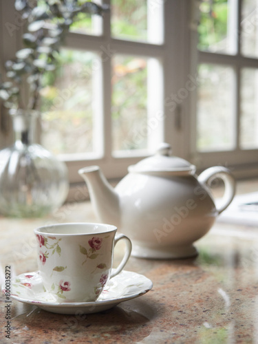 Close-up of porcelain tea cup and teapot on kitchen countertop indoors in a cozy cottage. Window out of focus in the background.