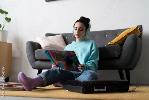 A young woman in a blue sweater examines a vinyl record, engaged in her passion for music, with a turntable and headphones nearby photo