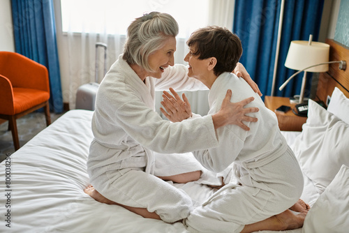 Two senior women enjoying a tender moment, sitting on a luxurious bed in a hotel.