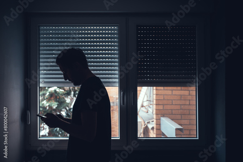 Depressed sad man using mobile phone in dark room by the window with shutters pulled down