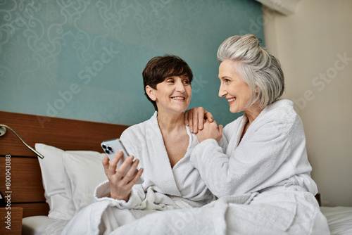 A senior lesbian couple in robes sitting closely on a bed, showing love and tenderness.