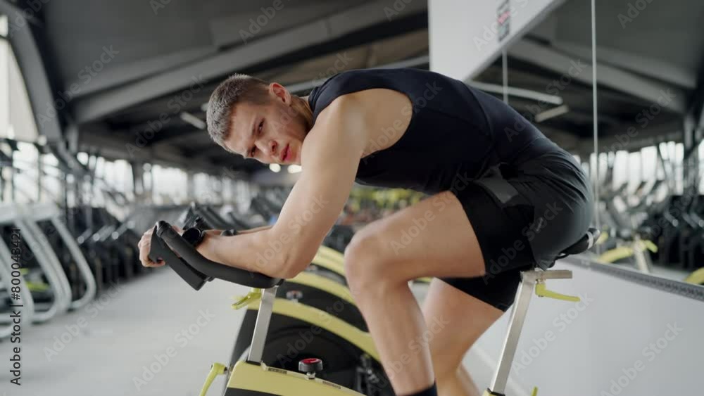A young male athlete in sports attire intensely working out on a stationary bike at a modern gym, displaying focus and determination. Focused Male Athlete During Intense Workout on Stationary Bike A young male athlete in sports attire intensely working out on a stationary bike at a modern gym, displaying focus and determination. Focused Male Athlete During Intense Workout on Stationary Bike
