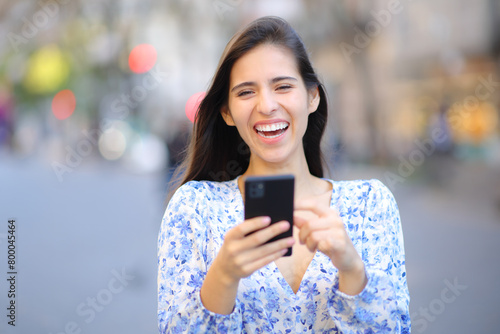 Happy woman laughing holding phone looking at you in the street