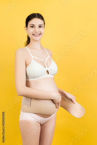 Portrait of pregnant woman in colorful underwear putting on a bandage at yellow background with copy space. Orthopedic abdominal support belt concept