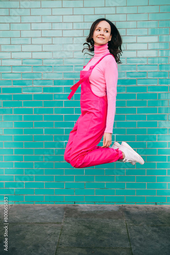 Smiling woman wearing pink bib overalls jumping in front of blue brick wall photo