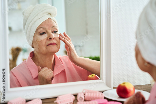 Senior woman wearing towel and applying face cream looking in mirror at home photo
