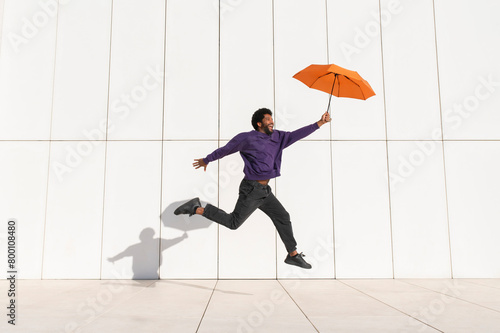 Cheerful man jumping with orange umbrella in front of white wall photo