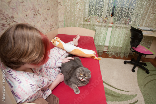 A girl in sleeping clothes sits on a bed and hugs her pet, a Scottish fold cat. An image about attitude and love for pets.