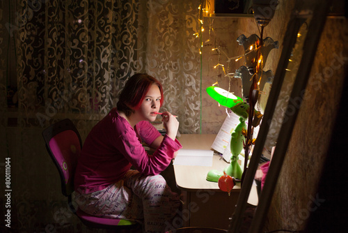 A schoolgirl girl with red hair sits at a table with a table lamp and a luminous garland, reads a book, does her homework, in a dark room in the evening. Image about studying at home.