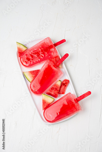 Aerial view of three watermelon lollies on white plate with pieces of watermelon, white background, vertical, with copy space