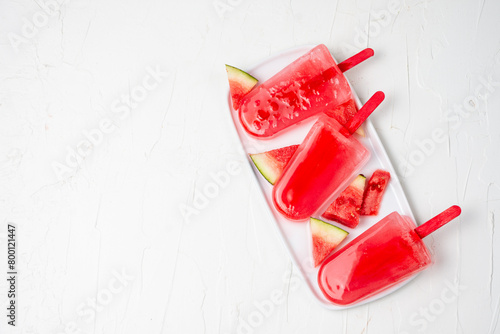 Top view of three watermelon lollies on white plate with watermelon pieces, white background, horizontal, with copy space