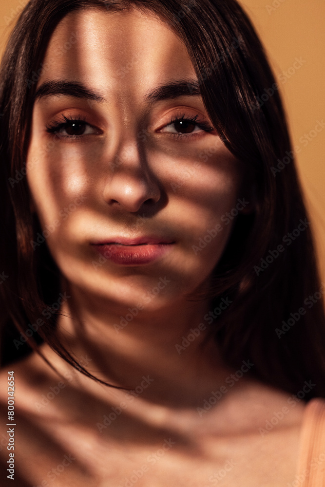A close-up portrait of a thoughtful female teenager in the warm lighting of the room. A sweet beautiful girl with a shadow of leaves on her face.