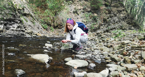 Tourist drinks from river. Close-up of girl traveler drinking water from mountain river. Stones, forest view, small pond. Danger of bacterial infection and diarrhea due to drinking unboiled water