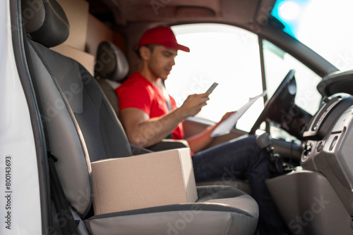 Courier man checking customer address using cellphone, sitting at drivers seat in van, focus on parcel nearby © Home-stock
