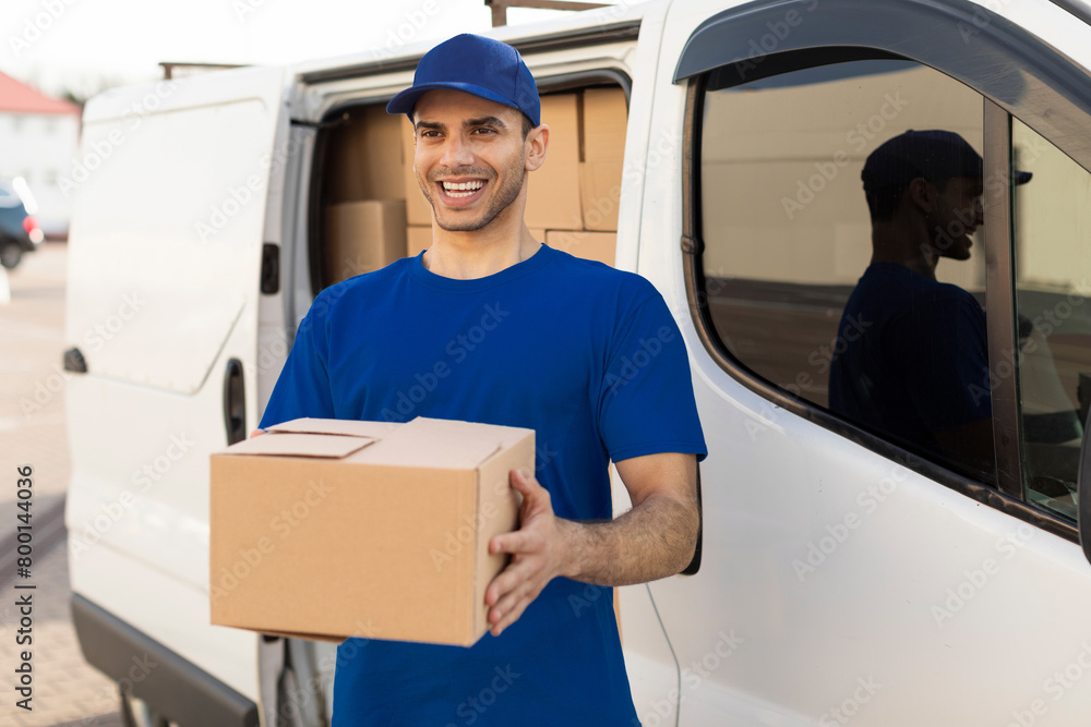 Mail service and shipment concept. Happy delivery man in uniform with parcel box looking and smiling at camera standing outdoors near van Mail service and shipment concept. Happy delivery man in uniform with parcel box looking and smiling at camera standing outdoors near van