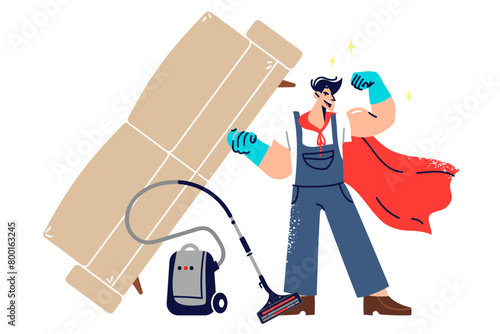Man janitor superhero demonstrates strength by lifting heavy sofa, standing next to vacuum cleaner