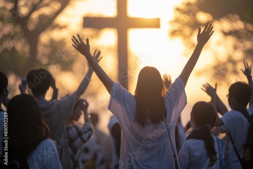 A crowd of various people are raising their two hands to the air to praise the Lord and  gathered around a wooden cross settled at a mound among the cloudy sky. Religion and faith in God.