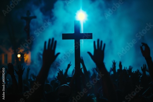 A crowd of various people are raising their two hands to the air to praise the Lord and gathered around a wooden cross settled at a mound among the cloudy sky. Religion and faith in God.