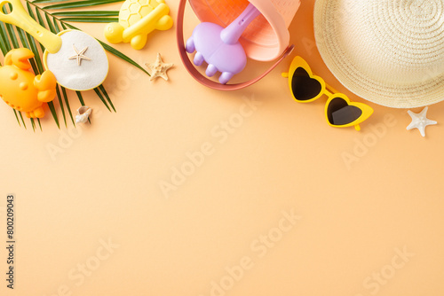 Child-friendly beach vacation scene. Flat lay top view composition featuring sand play tools, sunhat, shades, seashells, starfish, palm leaf on pastel orange surface. Text space available