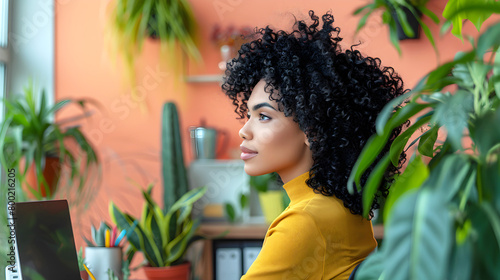 Young african american woman with curly hair sitting at desk in peach office, surrounded by vibrant and neatly arranged workspace photo