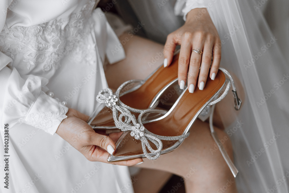 Fototapeta premium A woman is holding a pair of high heels with a bow on them. The shoes are silver and sparkly, and the woman is wearing a white dress. Concept of elegance and sophistication