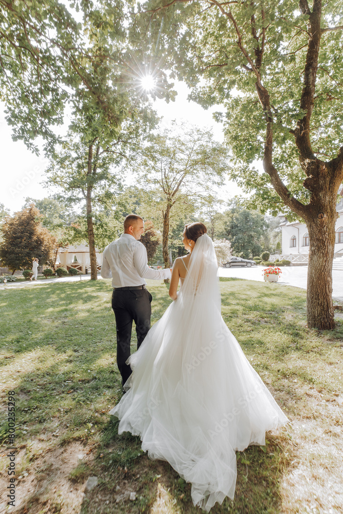 A bride and groom are walking together in a park. The bride is wearing a white dress and the groom is wearing a white shirt. The sun is shining brightly, creating a warm and happy atmosphere