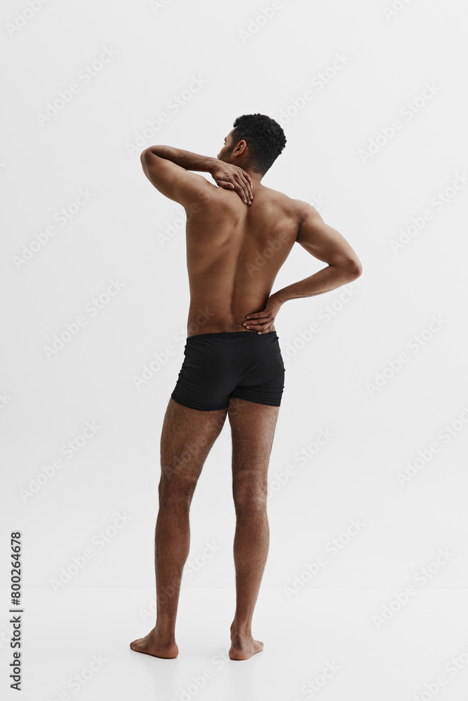 Fototapeta premium Rear view of young African-American shirtless man holding neck and back, standing in underwear isolated on white background. Health care. Concept of male beauty, sport, body care, health, fitness