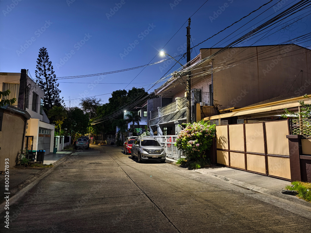 Las Pinas, Metro Manila, Philippines - April 28, 2024: Evening scene on a typical suburban street in BF Resort Village with houses and parked cars under a vibrant sunset sky. Las Pinas, Metro Manila, Philippines - April 28, 2024: Evening scene on a typical suburban street in BF Resort Village with houses and parked cars under a vibrant sunset sky.