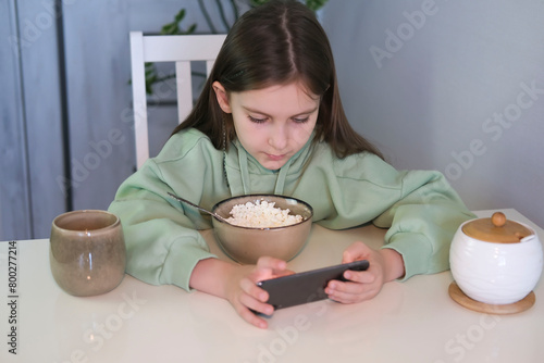 Teenage girl has breakfast in kitchen, uses mobile phone while having breakfast on table. Many children do not want to eat unless they are watching a mobile device or TV.