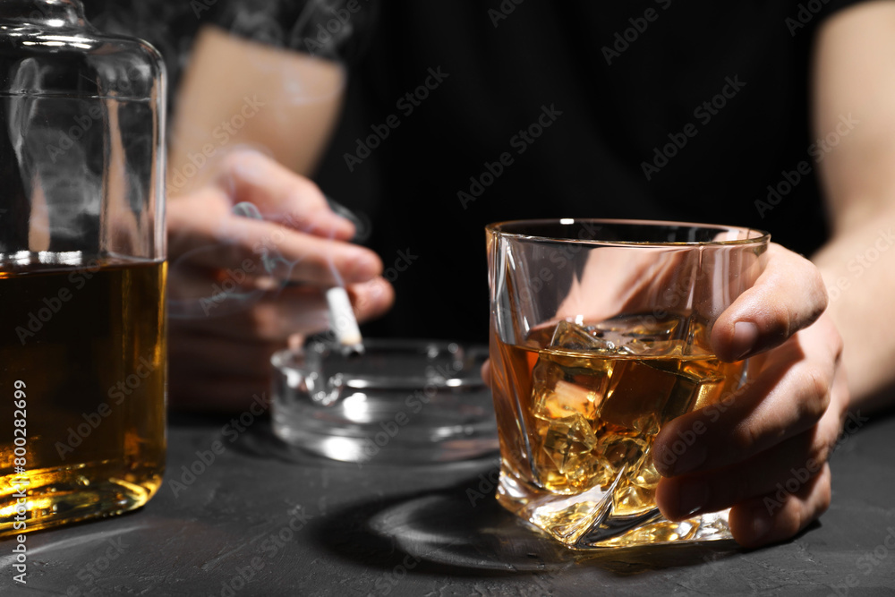Alcohol addiction. Man with glass of whiskey and smoldering cigarette at dark textured table, selective focus Alcohol addiction. Man with glass of whiskey and smoldering cigarette at dark textured table, selective focus