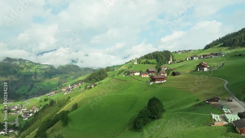 Flying above lush greenery towards a church on the steep slopes of the village of La Va, South Tyrol, Italy. Aerial footage  with cloud covered mountains in the background. LuPa Creative.