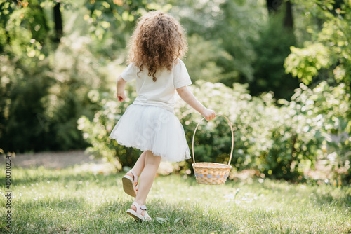 Easter egg hunt. Girl child Wearing Bunny Ears Running To Pick Up Egg In Garden. Easter tradition. Baby with basket full of colorful eggs.