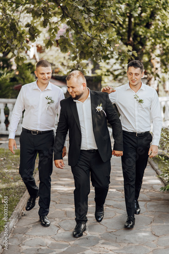 Three men in suits walking down a path with flowers on their collars. Scene is lighthearted and celebratory, as the men are dressed in formal attire and appear to be enjoying themselves
