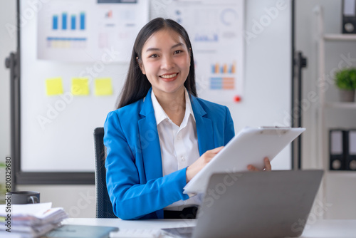 Beautiful young Asian business woman manager or company worker holding accounting document, checking financial data or marketing report working in office with laptop.
