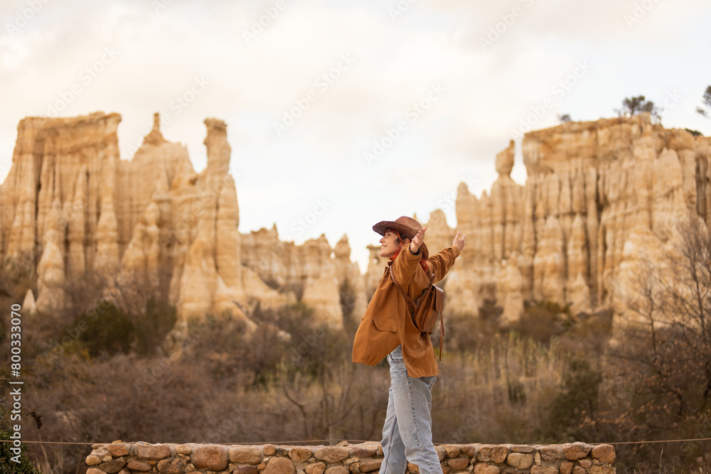 A woman with red hair and a cowboy hat stands in front of a mountain. She is wearing a brown coat and a hat