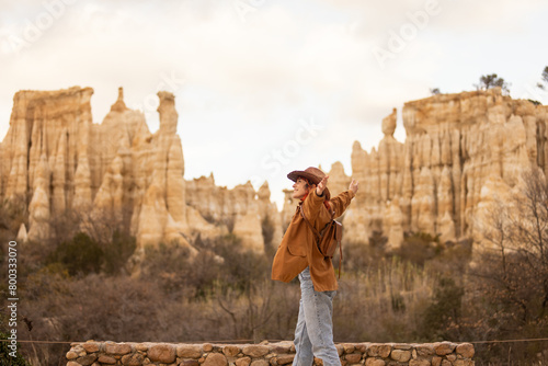 A woman with red hair and a cowboy hat stands in front of a mountain. She is wearing a brown coat and a hat