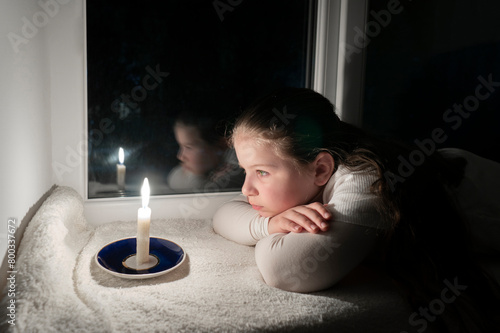 Blackout. A girl in a dark room lies on the windowsill near the window and looks at a burning candle