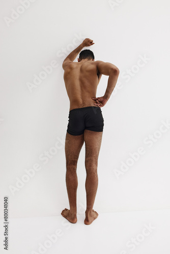 Young African-American man with relief strong back, muscular body standing shirtless in black boxers isolated on white background. Concept of male beauty, sport, body care, health, fitness