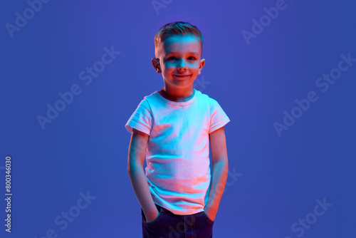 Cheerful smiling little boy dressed casual attire posing looking at camera in mixed neon light against blue background. Concept of human emotions, fashion and style, beauty, back to school. Ad
