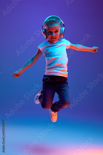 Schoolboy in casual attire jumping of joy while dancing during listening music in headphones in mixed neon light against blue background. Concept of music, dance, fashion and style, technology. Ad